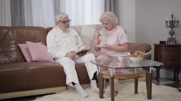 Portrait of mature Caucasian man reading book for his beautiful wife in the evening. Positive spouses drinking tea and resting together indoors. Happiness, love, leisure, lifestyle. — Stock Video