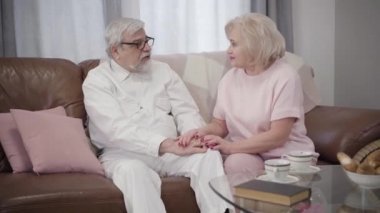 Elderly Caucasian couple sitting on couch holding hands and talking. Married mature spouses chatting at home in the evening. Happiness, bonding, lifestyle, eternal love.