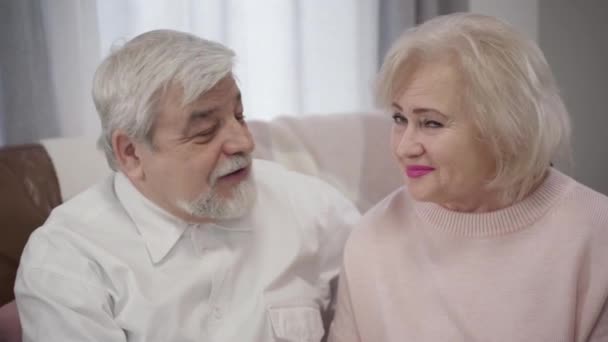 Primer plano de ancianos caucásicos hombre y mujer mirándose, girando hacia la cámara y sonriendo. Retrato de viejos esposos felices en el interior. Romance, amor eterno, estilo de vida . — Vídeo de stock