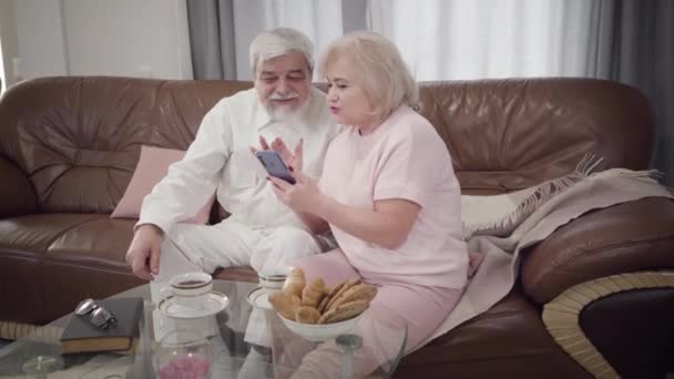 Portret van volwassen blanke man en vrouw met smartphone thuis. Moderne gepensioneerden zitten binnen op de bank te praten en te glimlachen. Geluk, vrije tijd, levensstijl. — Stockvideo