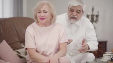 Portrait of irritated old Caucasian woman talking to husband. Elderly wife complaining to spouse, focus changing at senior man apologizing at the background. Lifestyle, conflict, arguing.
