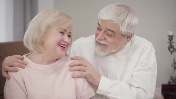 Elderly Caucasian couple talking to each other, looking at camera and smiling. Positive married retirees posing indoors. Love, happiness, lifestyle. — Stockvideo