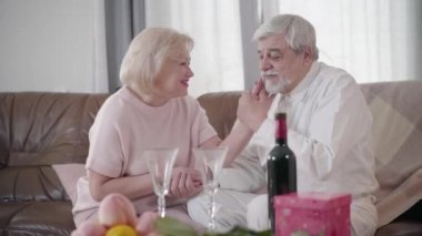 Side view of beautiful Caucasian elderly woman caressing husbands face and looking at him with love. Happy mature man kissing wifes hand and smiling. Romance, joy, eternal love concept.