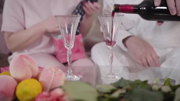 Κοντινό πλάνο ώριμων αρσενικών λευκών χεριών να ρίχνουν κρασί στα ποτήρια του κρασιού. Senior αγνώριστο παντρεμένο ζευγάρι γιορτάζει Αγίου Βαλεντίνου στο σπίτι. Γιορτή, γιορτή, αιώνια αγάπη. — Αρχείο Βίντεο