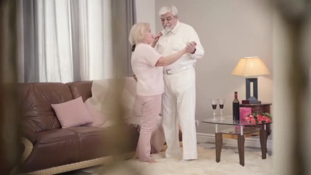 Largo tiro de madura pareja casada caucásica hablando y bailando en casa. Felices jubilados mayores celebrando el Día de San Valentín en el interior. Amor eterno de los esposos elegantes. Unidad, vinculación, estilo de vida . — Vídeos de Stock