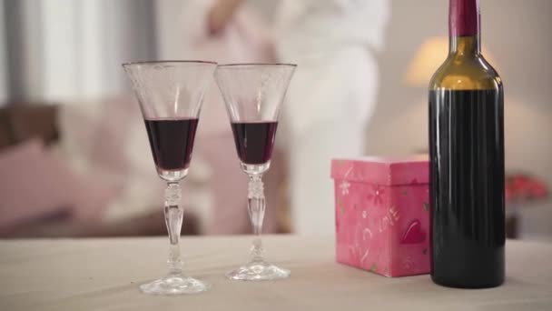 Közelkép két borosüvegről és üvegről, amint az asztalon állnak ajándékdobozzal. Homályos elegáns, fehér idős pár táncol a háttérben. Szerelem, romantika, Szent Valentin nap. — Stock videók