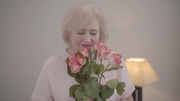 Primer plano retrato de hermosa mujer madura caucásica oliendo ramo de rosas, mirando a la cámara y sonriendo. Feliz jubilada senior posando con regalo de San Valentín en el interior . — Vídeo de stock