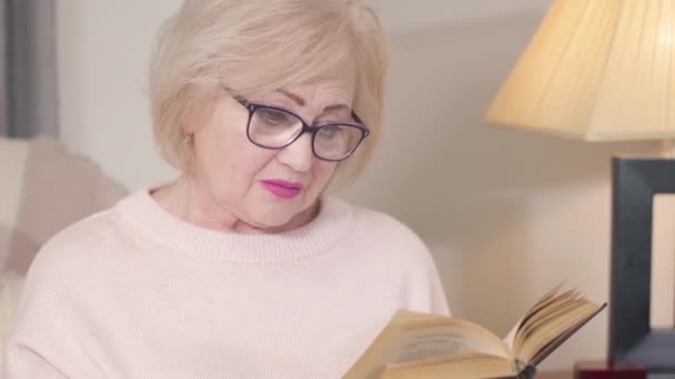 Nahaufnahme Porträt einer konzentrierten kaukasischen alten Frau, die Buch liest, sich der Kamera zuwendet und lächelt. Reife Frau mit Brille posiert zu Hause. Freizeit, Hobby, Intelligenz, Lebensstil. — Stockvideo