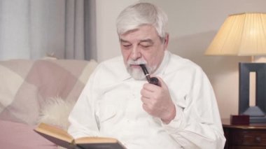 Beyaz saçlı beyaz adamın portresi pipo içiyor ve evde kitap okuyor. Olgun, ciddi bir adam evde sakin bir akşam geçiriyor. Boş zaman, hobi, yaşam tarzı..