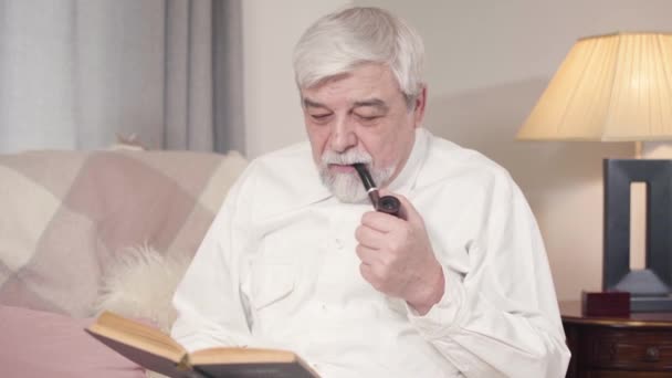 Portrait d'homme caucasien aux cheveux gris fumant de la pipe et lisant un livre à la maison. Mature homme sérieux passer la soirée tranquille à l'intérieur. Loisirs, passe temps, mode de vie . — Video
