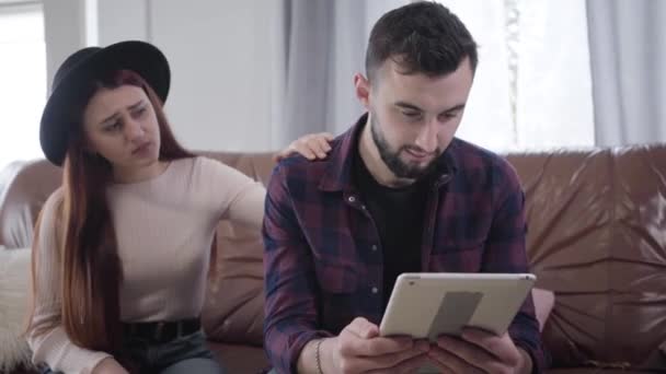 Junge kaukasische Mann mit Tablet als seine schöne traurige Freundin oder Frau, die mit ihm im Hintergrund spricht. Ehemann oder Freund ignorieren Ehepartner zu Hause. Ehe, Problem. Mitteilung. — Stockvideo