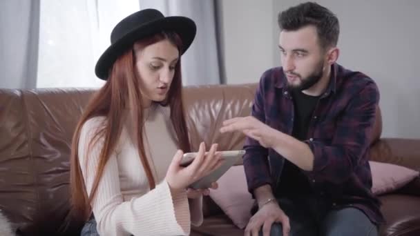 Joven linda mujer caucásica irritante como su marido o novio explicando cómo utilizar la tableta. Chica elegante en sombrero discutiendo con el cónyuge en casa. Estilo de vida, problemas de comunicación . — Vídeo de stock