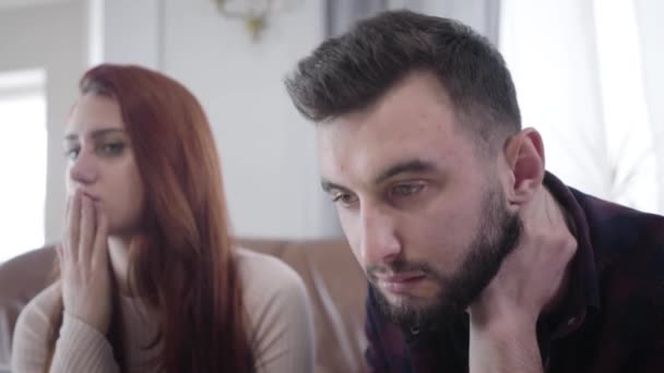 Närbild av unga skäggiga kaukasiska mannen med bruna ögon rörande ansikte nervöst som orolig vacker flickvän eller fru sitter i bakgrunden. Unga par väntar på psykologisk rådgivning. — Stockvideo