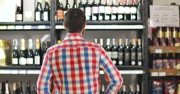 Vue arrière de l'homme qui se gratte la tête en regardant des étagères avec des bouteilles de vin. Client caucasien confus choisir une boisson dans un luxueux magasin de vin. Alcool, boisson, consumérisme, mode de vie . — Video