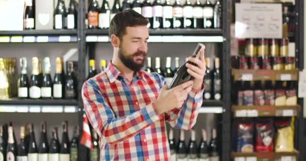 Jonge vrolijke blanke man kijkt naar label op wijnfles, knuffelen drankje, kijken naar camera en glimlachen. Tevreden klant koopt alcohol in luxe winkel. Lifestyle, geluk, drank. — Stockvideo