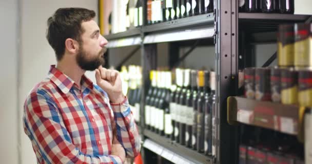 Vista lateral del hombre caucásico barbudo reflexivo mirando estantes con botellas de vino y la cabeza arañazos. Joven concentrado eligiendo alcohol en una tienda de lujo. Industria del alcohol, estilo de vida, riqueza . — Vídeos de Stock