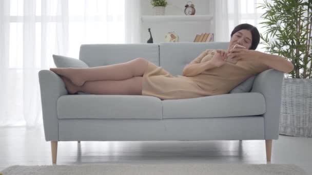 Relaxed Caucasian senior woman in elegant beige dress lying on sofa and talking on the phone. Happy middle-aged female retiree spending free time at home. Lifestyle, leisure, tranquility. Wide shot. — Stock Video