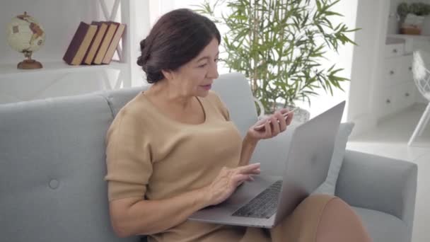 Femme caucasienne âgée confiante utilisant un ordinateur portable et un smartphone comme assise sur un canapé à l'intérieur. Brune réussie femme d'affaires d'âge moyen avec les yeux bruns travaillant à la maison. Mode de vie, succès, richesse . — Video