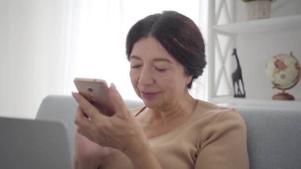 用智能手机拍摄的聪明的白人老年女性的特写。自信的中年退休人员敲击屏幕，在室内上网。生活方式、休闲、智力、现代技术 — 图库视频影像