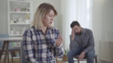 Depresyonda olan beyaz bir kadın, elinde alyansla yüzüne dokunuyor. Arka planda oturan stresli koca. Mutsuz çift içeride ayrılıyor. Boşanma, evlilik..