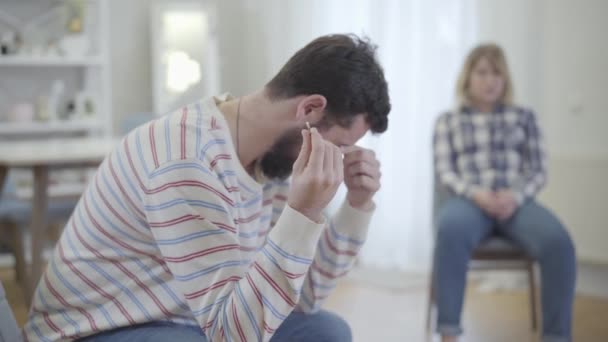 Vista lateral de un joven caucásico deprimido sosteniendo el anillo de bodas y frotando los ojos doloridos con la mano. Mujer rubia borrosa sentada al fondo. Los cónyuges rompen. Divorcio, problemas de relación . — Vídeos de Stock