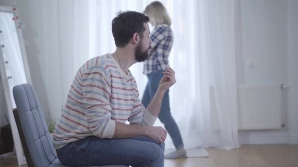 Young bearded Caucasian man looking at wedding ring in hand while nervous wife walking at the background. Stressed married couple breaking up. Divorce, relationship, conflict, breakup. — Stock Video