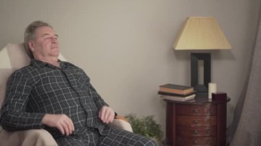 Sallanan sandalyede sallanan, gözlük takan ve kitap okuyan düşünceli beyaz adamın portresi. Yaşlı emekli içeride kitap okuyor. Hobi, yaşam tarzı, edebiyat, rahatlama.