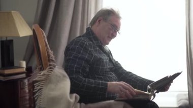 Olgun, beyaz, yaşlı bir adam elinde fotoğraf albümüyle pencerenin önünde oturuyor ve gülümsüyor. Yaşlı emeklinin kafa sallayıp öksürüşünün yan görüntüsü. Yaşlanma, hatıralar, yaşam tarzı.