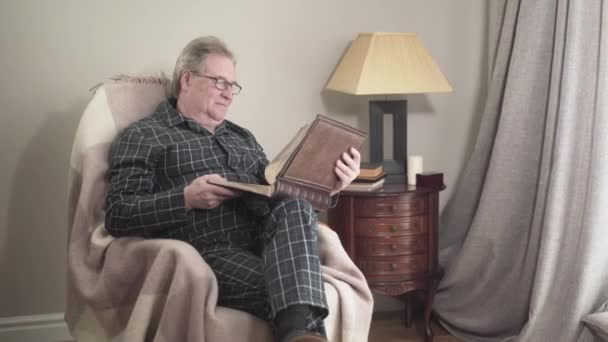 Alter kaukasischer Mann im Schlafanzug sitzt im Schaukelstuhl und betrachtet Fotos im Fotoalbum. Positiver Rentner mit Brille, der sich an Erinnerungen zu Hause erinnert. Lebensstil, Freizeit, Entspannung. — Stockvideo