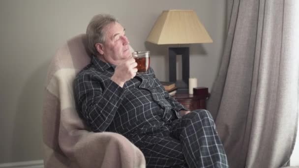 Portrét spokojeného dospělého bělocha mávajícího a ukazujícího palcem z okna. Senior šťastný důchodce sedí doma v houpacím křesle s šálkem čaje a usmívá se. Volný čas, odpočinek, životní styl. — Stock video