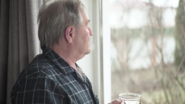 Side view portrait of thoughtful senior Caucasian man looking out the window and drinking tea indoors. Lonely male retiree waking up in the morning at home. Lifestyle, leisure, retirement, aging. — Stockvideo