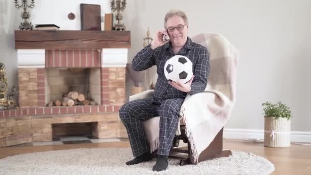 Веселый европеец, сидящий в кресле с футбольным мячом и разговаривающий по телефону. Портрет положительного пенсионера, увлеченного футбольным разговором с другом. Активный образ жизни, пенсия, жизнеспособность . — стоковое видео