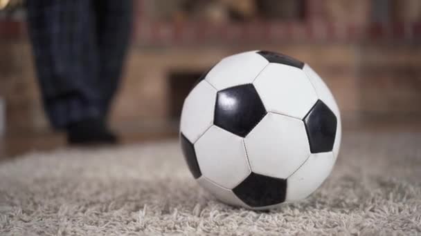 Primer plano de la pelota de fútbol tirada en el suelo en el interior. Pies masculinos en pijama golpeándolo y siguiéndolo. Concepto de estilo de vida saludable, actividad deportiva, vitalidad . — Vídeos de Stock