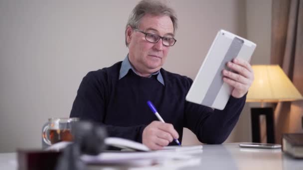 Senior Caucasian man writing at the table when having incoming video call. Mature male retiree in eyeglasses waving and talking at selfie camera. Lifestyle, modern technologies, communication. — Stockvideo