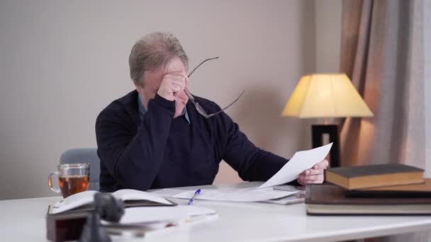 Portrait of tired senior Caucasian man taking off eyeglasses and putting away papers. Exhausted male author stuck without inspiration. Aging, creativity, lifestyle. — Stockvideo