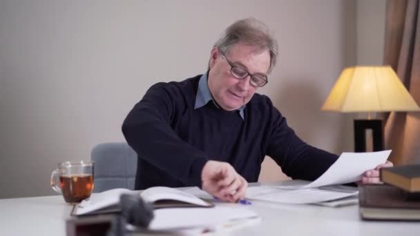 Felice uomo caucasico anziano che scrive sui giornali e sorride. Positivo autore attivo creando nuovo capolavoro all'interno. Ritratto di vecchio sorridente con gli occhiali che lavora. Stile di vita, letteratura, creatività . — Video Stock