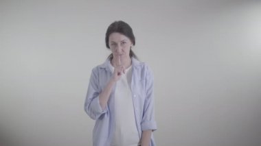 Yetişkin, beyaz bir kadının sessiz bir jest yapıp kameraya bakışının ortası. Beyaz arka planda poz veren esmer kadın. Sır saklama kavramı.