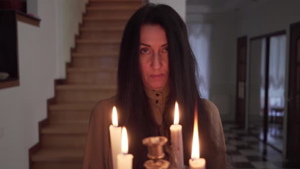 Portrait de femme brune mystérieuse regardant la caméra et partant avec des bougies allumées dans un chandelier. Gros plan de dame blanche effrayante à l'intérieur. Éerie, magie, paranormal, mystère . — Video