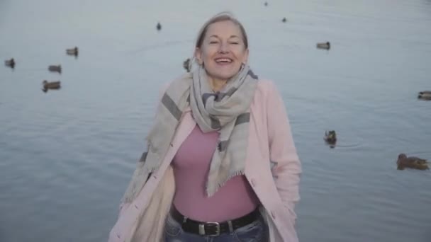 Foto en medio de una sonriente dama caucásica con abrigo rosa al fondo de patos volando y nadando en el río. Retrato de mujer adulta feliz disfrutando de un soleado día de otoño en la orilla del río. Ocio, estilo de vida . — Vídeo de stock