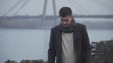 Nehrin arka planında kameraya doğru yürüyen üzgün, düşünceli beyaz adamın portresi. Sonbahar montlu esmer adam dışarıda yalnız bir gün geçiriyor. Yaşam tarzı, depresyon, düşünce.