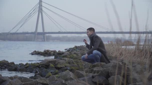 Pensativo joven caucásico sentado en la orilla del río y arrojando piedras al agua. Retrato de un chico guapo triste en depresión pasando el solitario día de otoño al aire libre. Estilo de vida, soledad, naturaleza . — Vídeo de stock