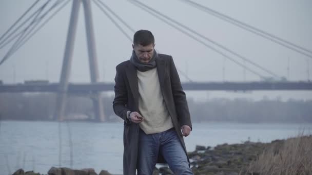 Middle shot of depressed Caucasian man in elegant coat and scarf walking on riverbank and throwing little rock in water. Angry young guy having problems. Loneliness, sadness, depression. — Stock Video