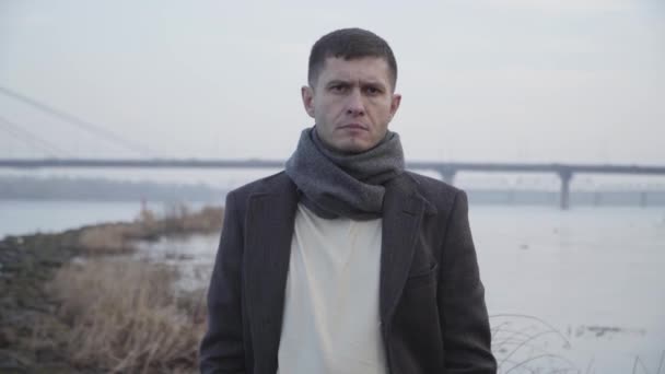 Retrato del infeliz hombre caucásico posando en el fondo del río y el puente de la ciudad. Joven trastornado mirando la cámara y suspirando. Problemas, estrés, depresión . — Vídeo de stock