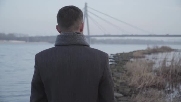 Pohled zezadu na elegantního bělocha stojícího na břehu řeky a dívajícího se na městský most v pozadí. Osamělý bruneta chlap odpočívá v parku na podzim. Životní styl, volný čas, samotu. — Stock video