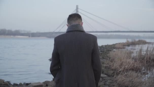 Middle shot of lonely Caucasian man walking along rocky riverbank and thinking. Back view of young guy in elegant coat strolling on grey autumn day outdoors. Loneliness, lifestyle, nature. — Stock Video