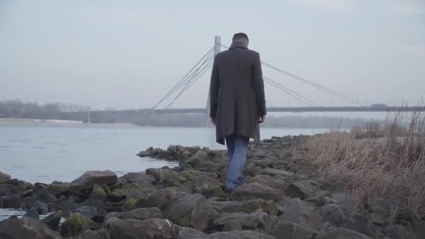 Ampia ripresa di uomo adulto caucasico passeggiare sulla riva rocciosa del fiume il giorno d'autunno. Un tipo triste e solitario che passa del tempo all'aperto da solo. Natura, tempo libero, solitudine . — Video Stock