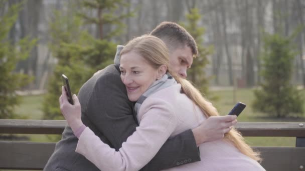 Side view van knuffelen Kaukasisch volwassen echtpaar met behulp van smartphones. Blond vrouw en brunette man met behulp van sociale media op een date buiten. Apparaatverslaving, sociale communicatie, levensstijl. — Stockvideo