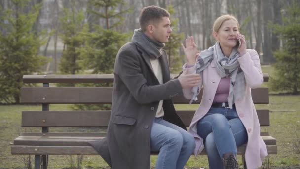 Mujer rubia caucásica adulta hablando por teléfono y riendo como un hombre más joven tratando de atraer su atención. Retrato de esposa o novia ignorando a un chico cariñoso en una cita. Molestia, irritación . — Vídeos de Stock