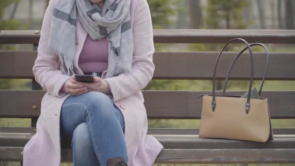 Unerkennbare Kaukasierin, die draußen auf einer Bank sitzt, während eine männliche Hand ihre Handtasche stiehlt. Unachtsame elegante Dame im rosafarbenen Mantel verliert ihre Handtasche, als sie soziale Medien im Smartphone nutzt. Stadtleben, Kriminalität. — Stockvideo