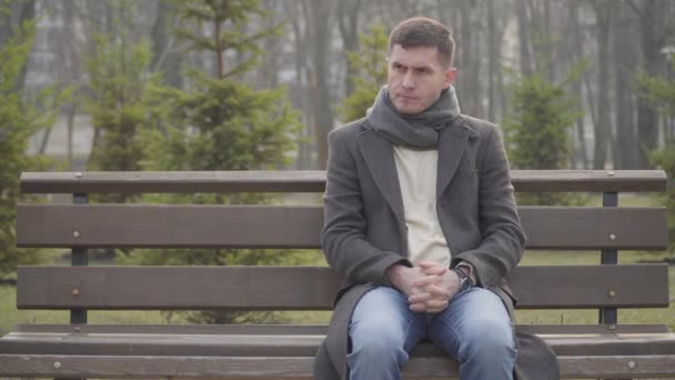 Jeune homme nerveux caucasien assis sur un banc dans le parc de la ville et vérifiant l'heure de quart. Portrait d'un mec brune impatient qui attend à l'extérieur le jour ensoleillé d'automne. Mode de vie, réunions, ponctualité . — Video